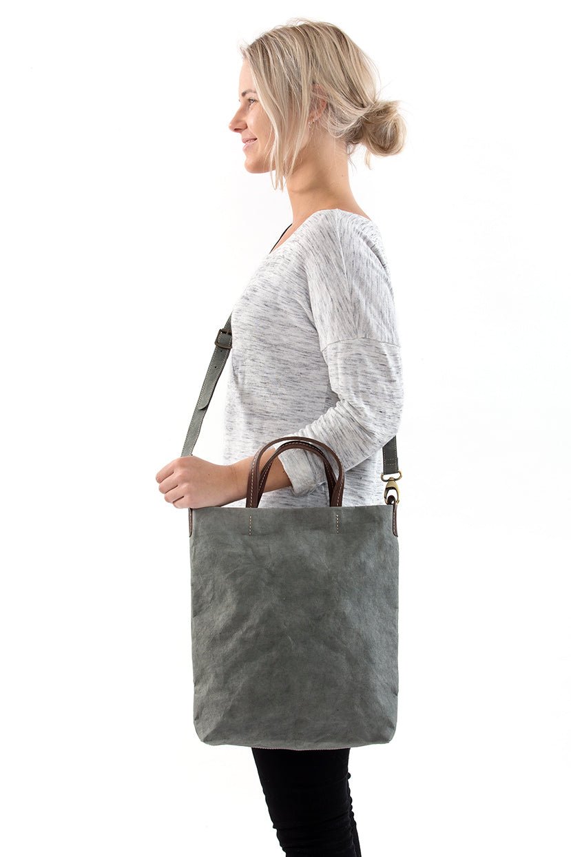 A blonde woman is shown from the side wearing casual clothing. She wears a crossbody washable paper tote bag with top handles. It is dark grey in colour.