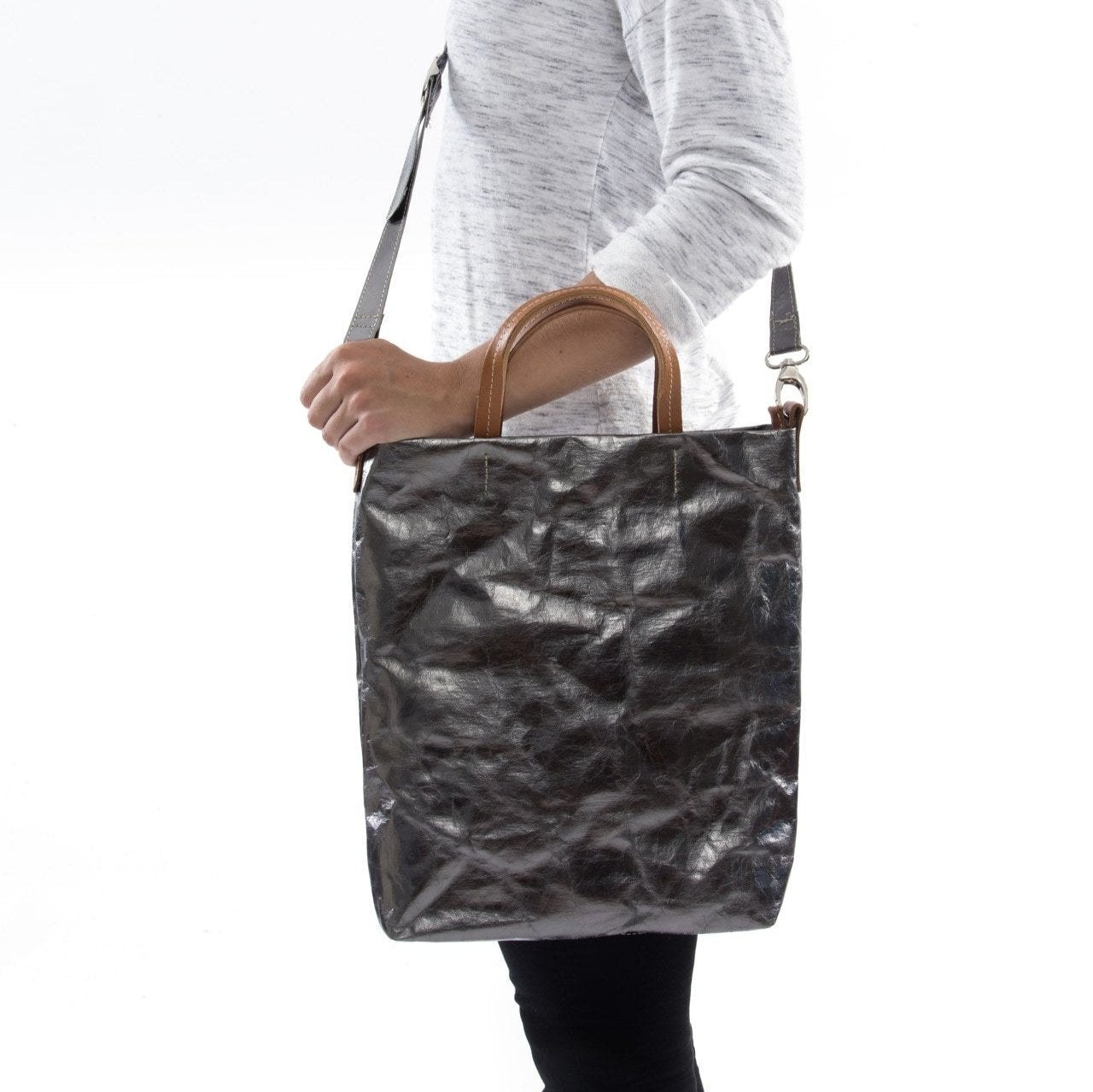 A blonde woman is shown from the side wearing casual clothing. She wears a crossbody washable paper tote bag with top handles. It is metallic pewter in colour.