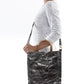 A blonde woman is shown from the side wearing casual clothing. She wears a crossbody washable paper tote bag with top handles. It is metallic pewter in colour.