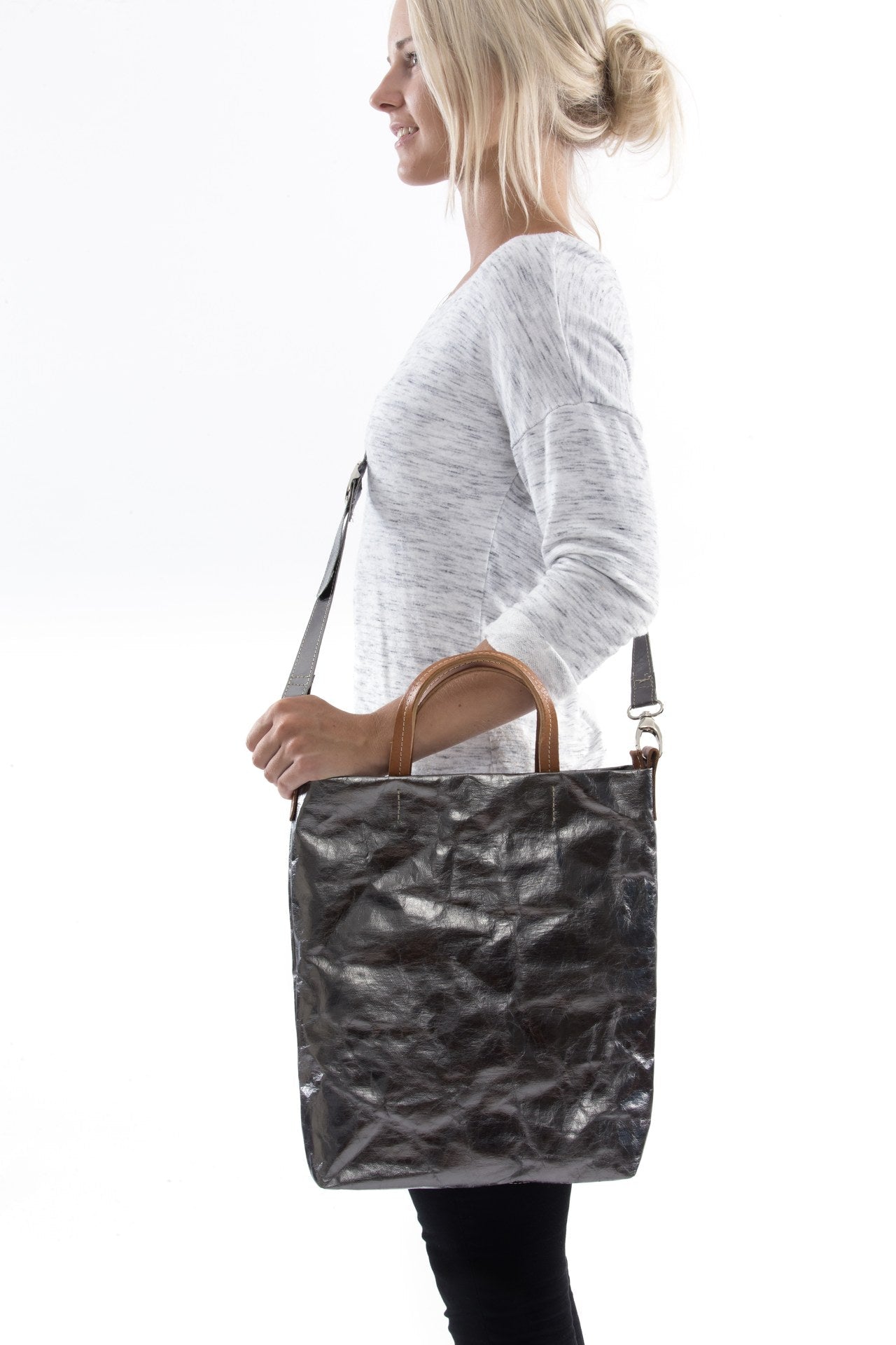 A blonde woman is shown from the side wearing casual clothing. She wears a crossbody washable paper tote bag with top handles. It is metallic pewter in colour.