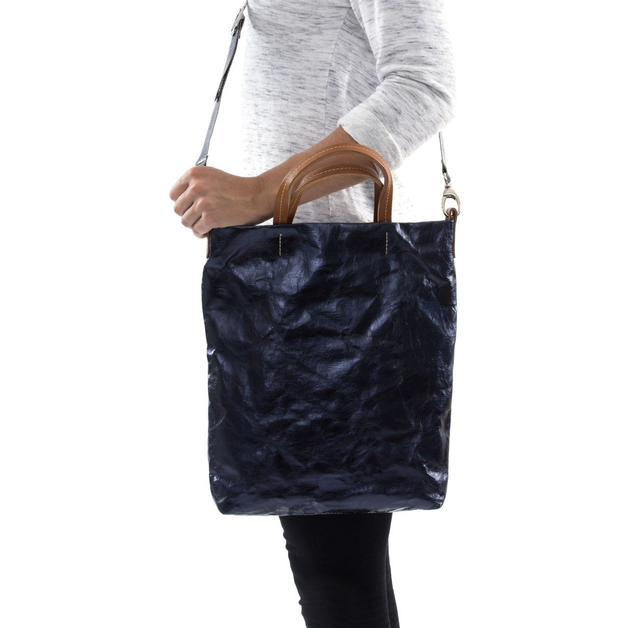 A blonde woman is shown from the side wearing casual clothing. She wears a crossbody washable paper tote bag with top handles. It is metallic navy in colour.