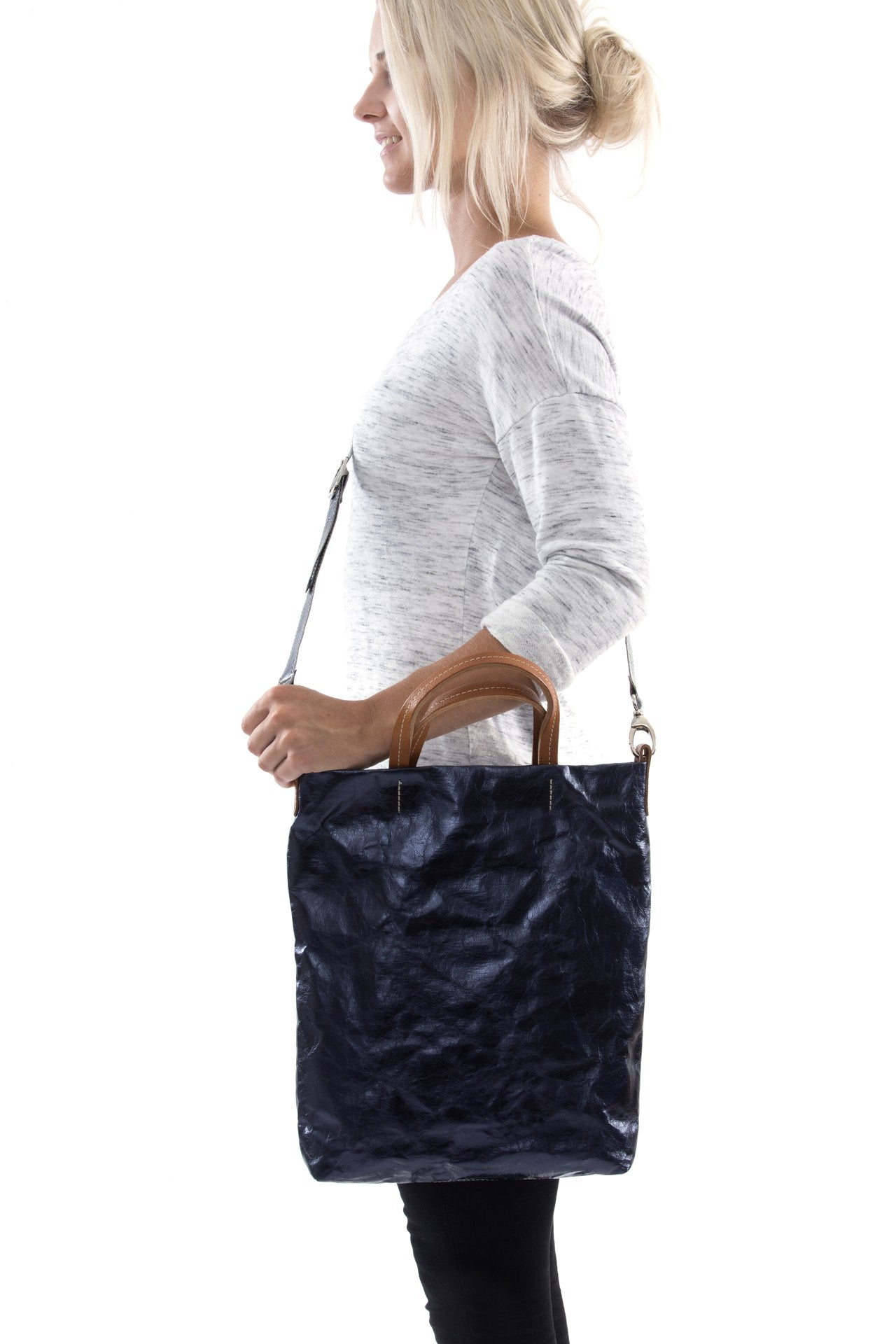 A blonde woman is shown from the side wearing casual clothing. She wears a crossbody washable paper tote bag with top handles. It is metallic navy in colour.
