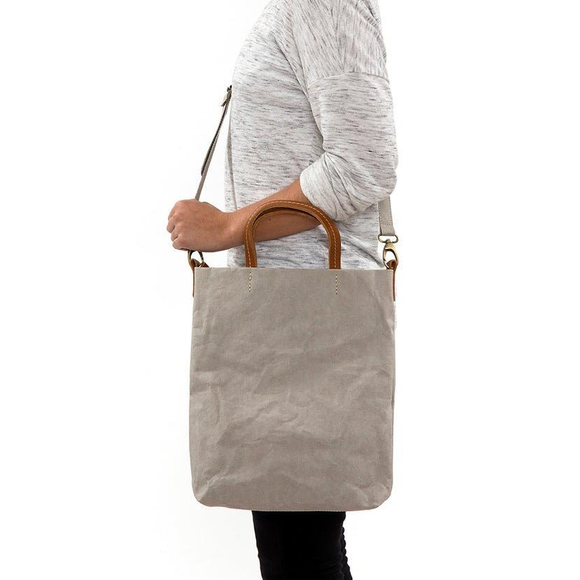 A blonde woman is shown from the side wearing casual clothing. She wears a crossbody washable paper tote bag with top handles. It is pale grey in colour.