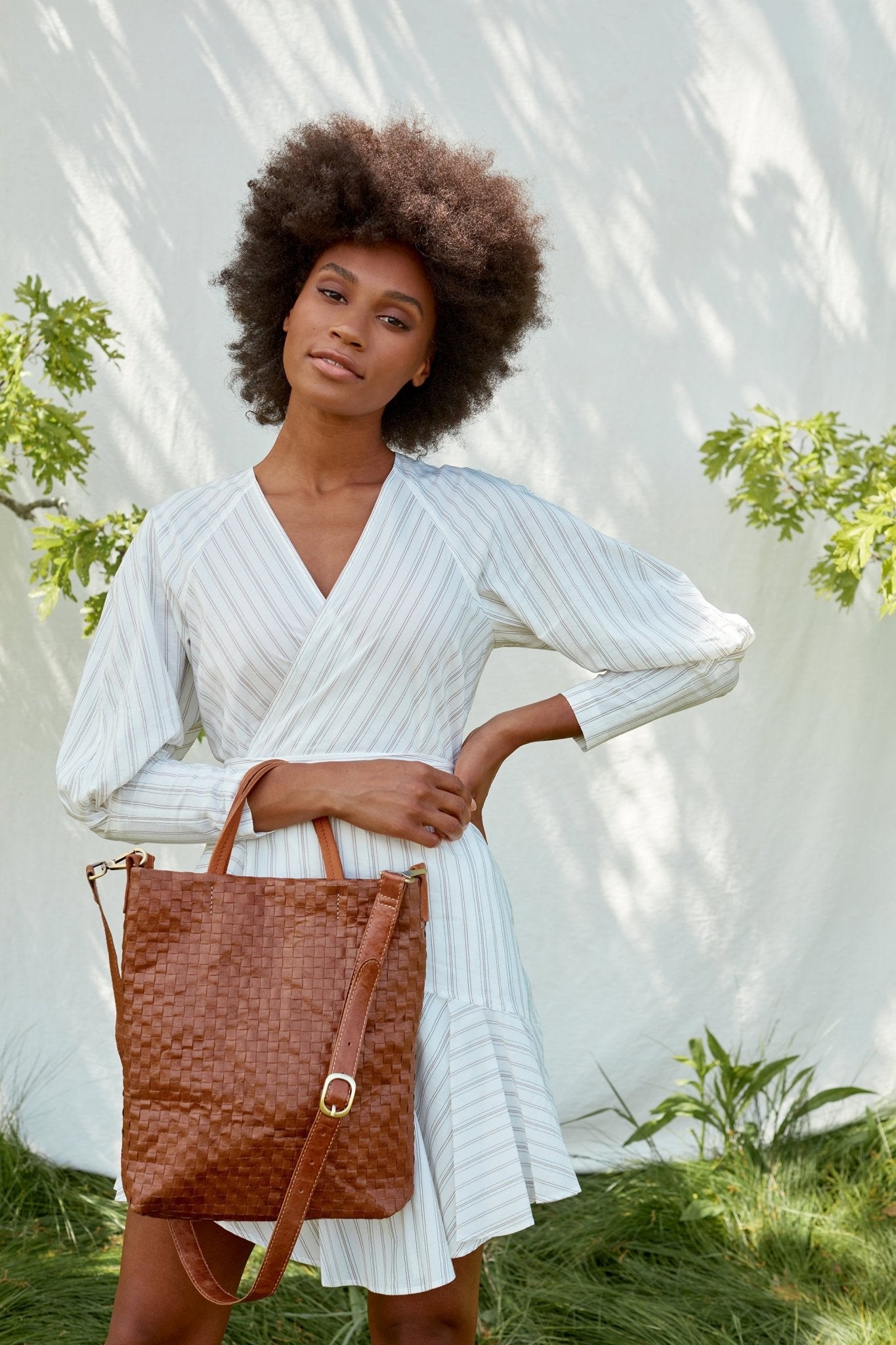 A woman is shown outdoors amidst foliage, in front of a linen backdrop. She wears a white dress and carries a brown woven washable paper tote bag over one arm.