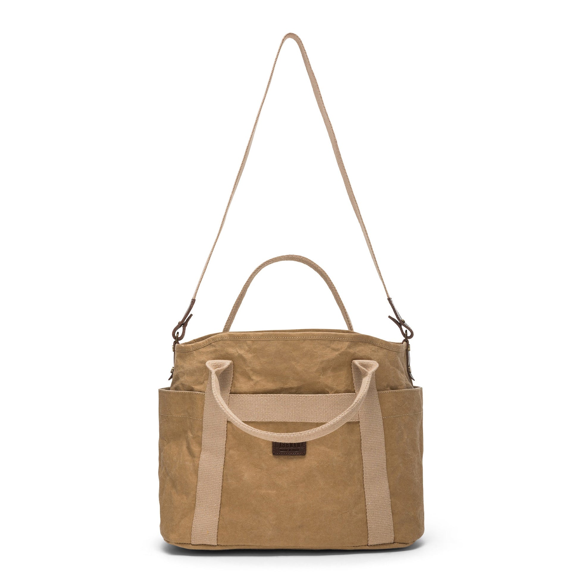 A large washable paper shopper bag is shown from a front angle. It features a long canvas shoulder strap and two top handles. It has an external pocket running the length of the bag, and the UASHMAMA logo stamped in the front. It is shown in a natural pale tan colour.