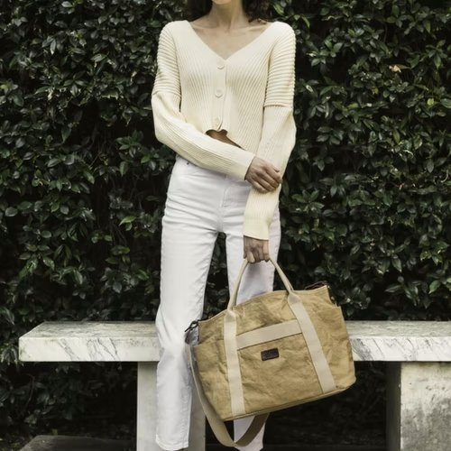 A woman is shown wearing a cream cardigan and white jeans, outdoors. She holds a washable paper shopper bag in a sandy colour in her left hand.