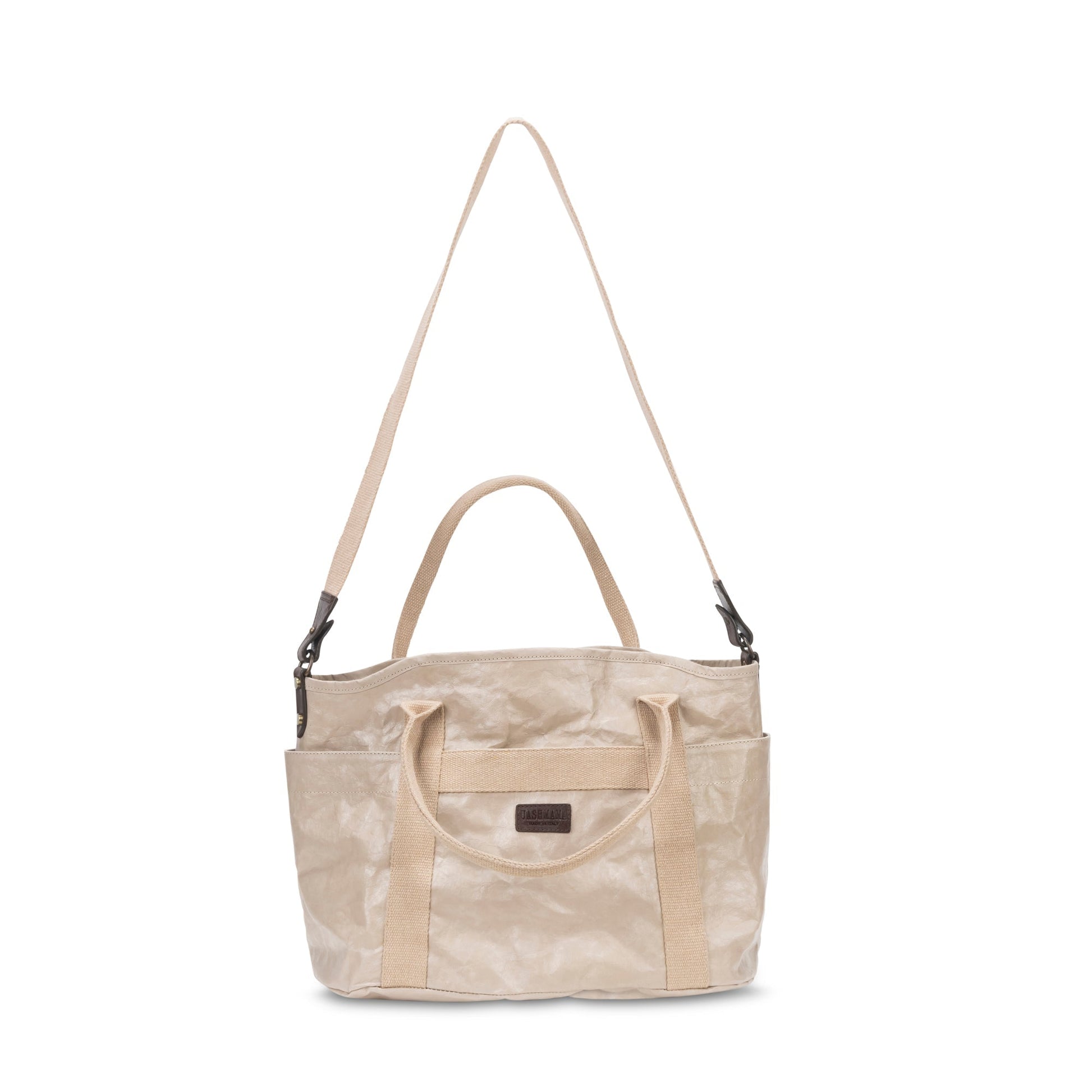A large washable paper shopper bag is shown from a front angle. It features a long canvas shoulder strap and two top handles. It has an external pocket running the length of the bag, and the UASHMAMA logo stamped in the front. It is shown in a cream colour.