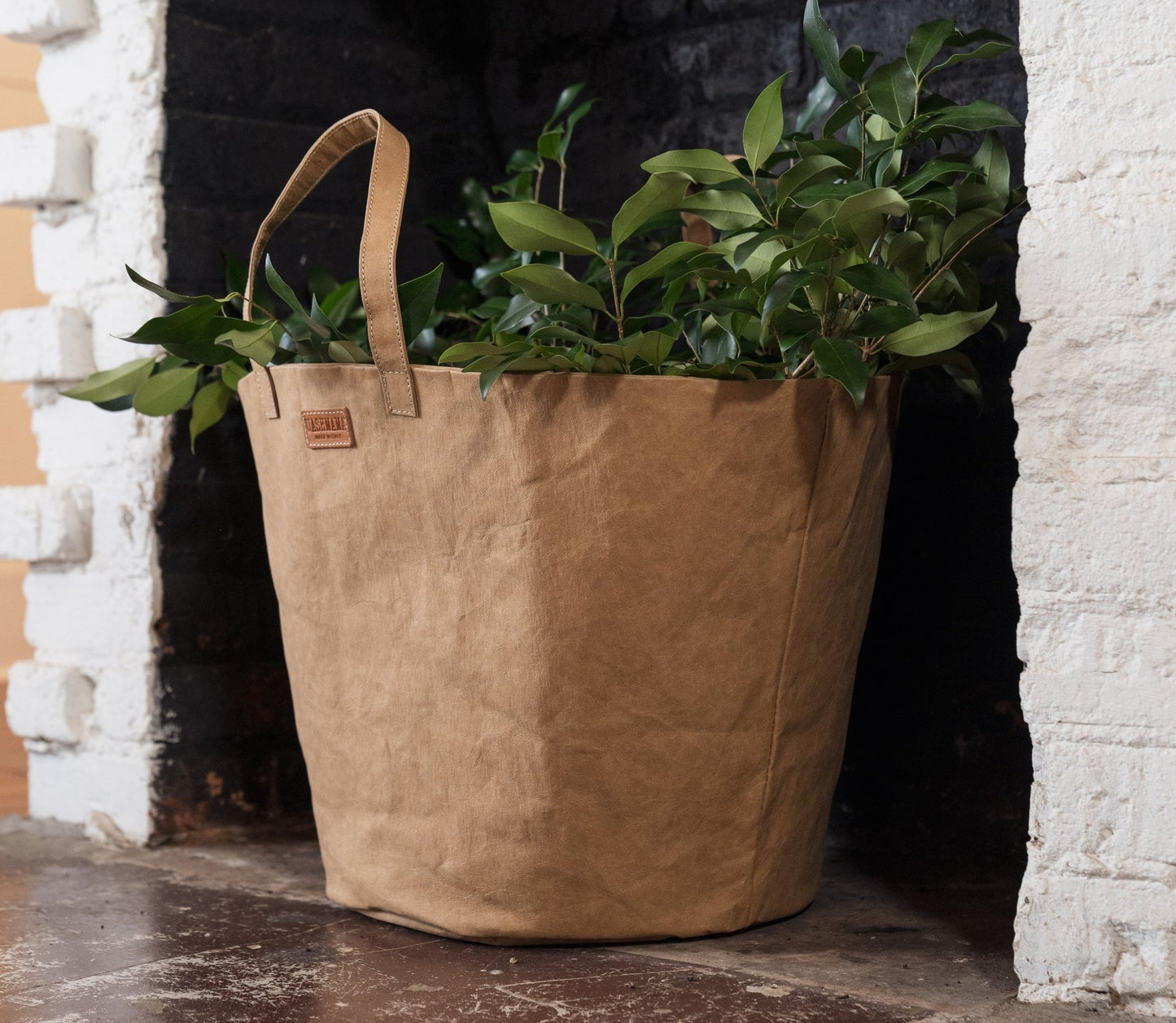 A washable paper basket with two top handles is shown in a fireplace, holding foliage. It is shown in a tan colour. 