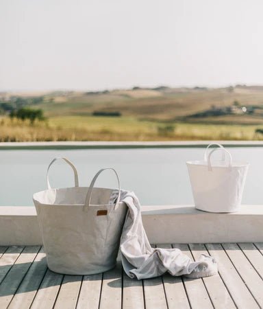 Two washable paper baskets sit outdoors on decking. The one in the front is shown in a cream colour, with a towel draped out of it. The one in the rear is smaller and shown in white.