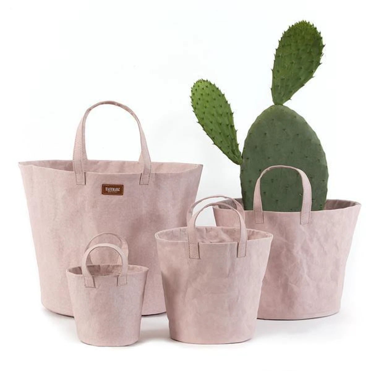 A selection of washable paper baskets are shown stacked in one another, in five varying sizes. They are shown in a pale pink colour.Four individual washable paper baskets are shown in varying sizes. The one at right contains a large cactus, and they are shown in a pale pink colour.