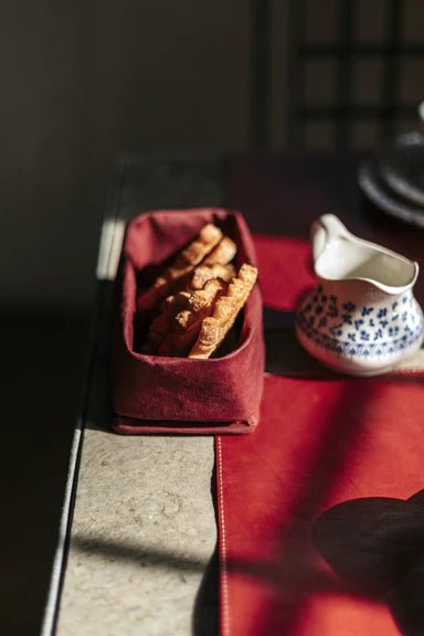 A table top shows a red washable paper tray containing sliced bread. A porcelain jug sits at right on top of a washable paper table runner.