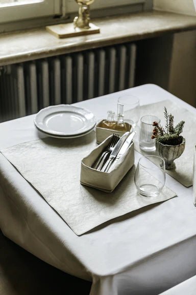 A table is set for two, with washable paper placemats and a washable paper small oil and vinegar holder. In the foreground sits a washable paper tray in grey holding cutlery.