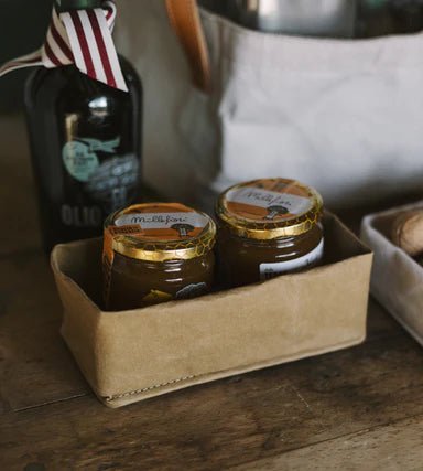 A tan washable paper tray sits atop a wooden table containing marmalade and jam. A bottle of olive oil sits in the background.
