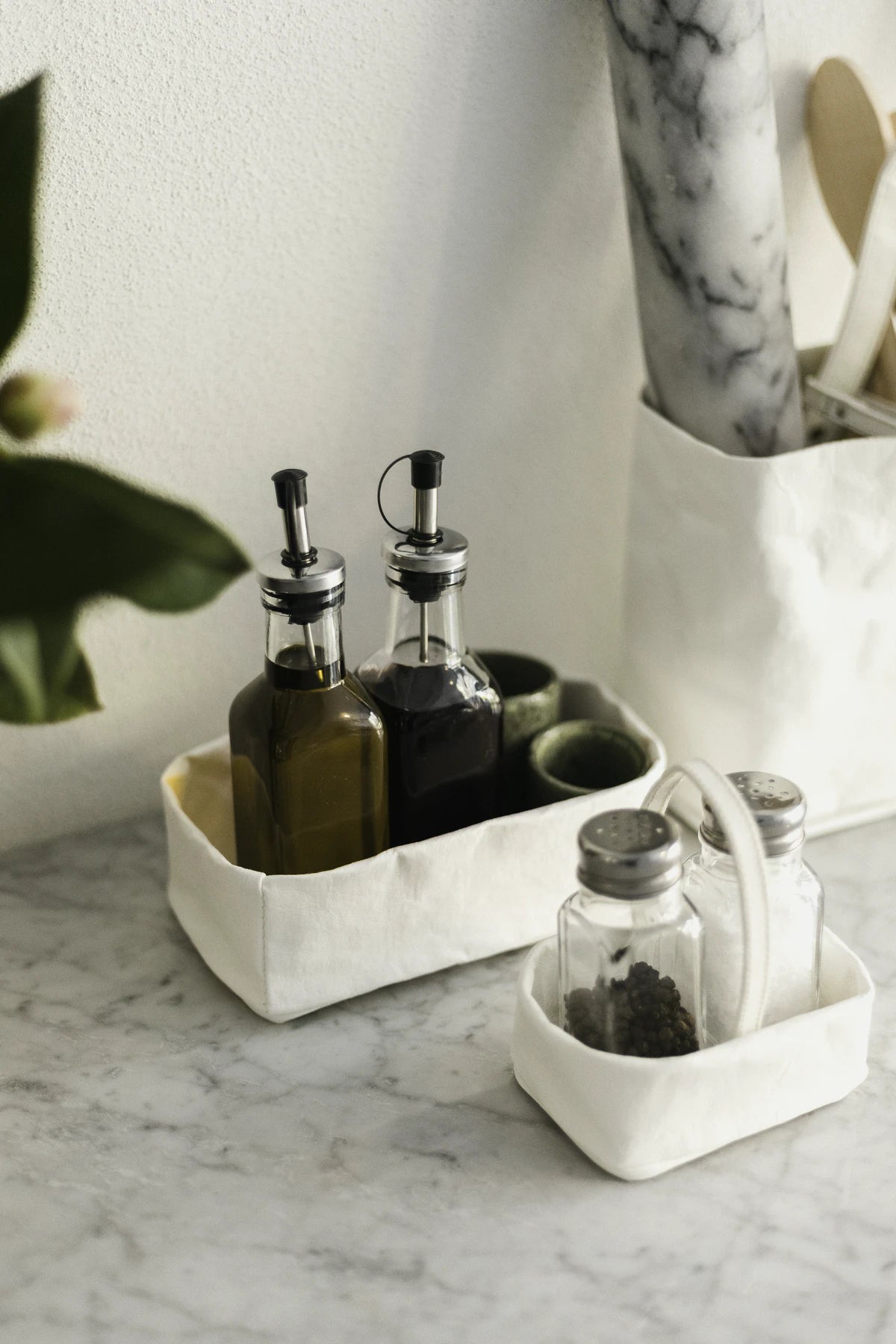 A washable paper tray is shown holding a bottle of oil and a bottle of vinegar. It sits atop a marble sink, and a small washable paper utensil holder sits in front containing salt and pepper.
