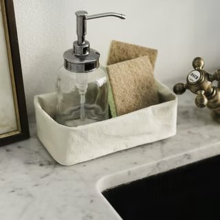 A washable paper tray is shown in cream, containing dish sponges and a soap dispenser. It sits on the surface of a marble sink.