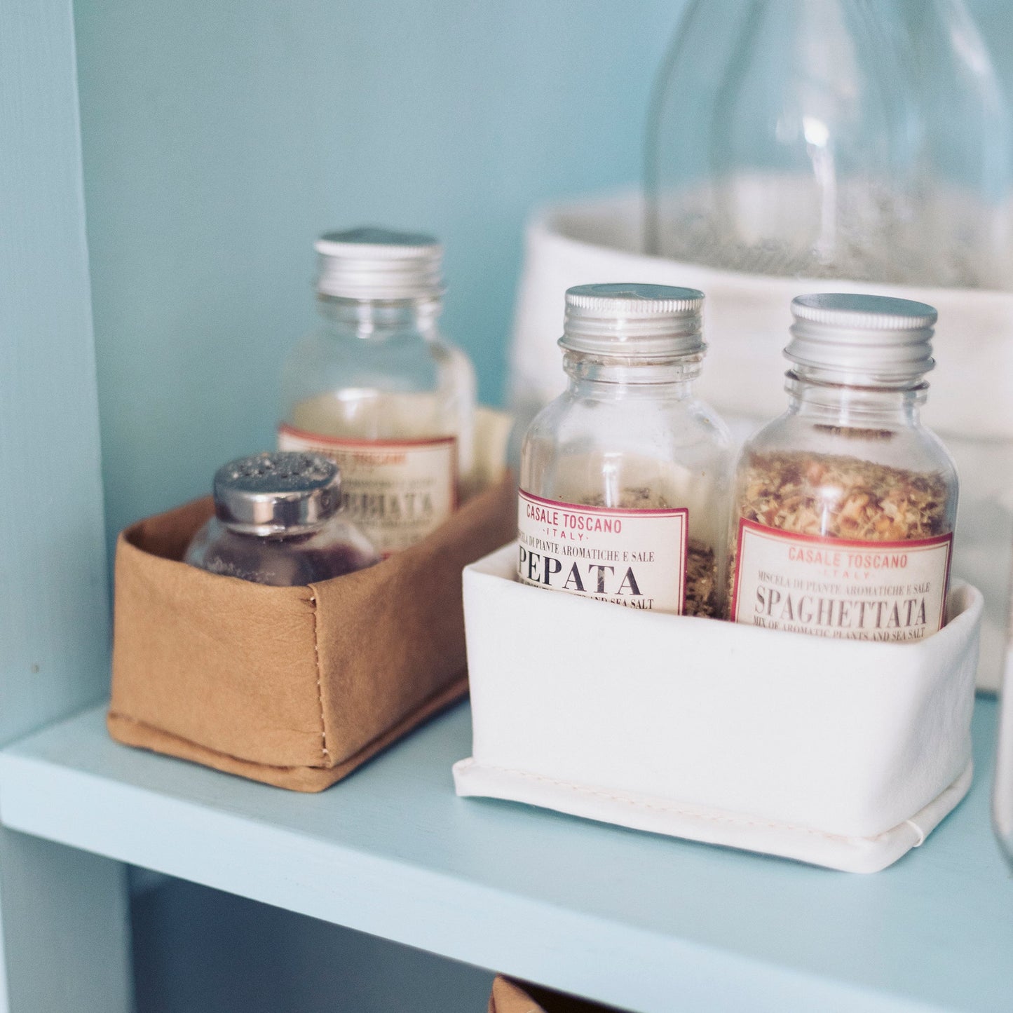 Three washable paper trays in varying sizes sit on a kitchen shelf. The two in the foreground are small, containing spices. The one at left is tan in colour and the one at right is white.