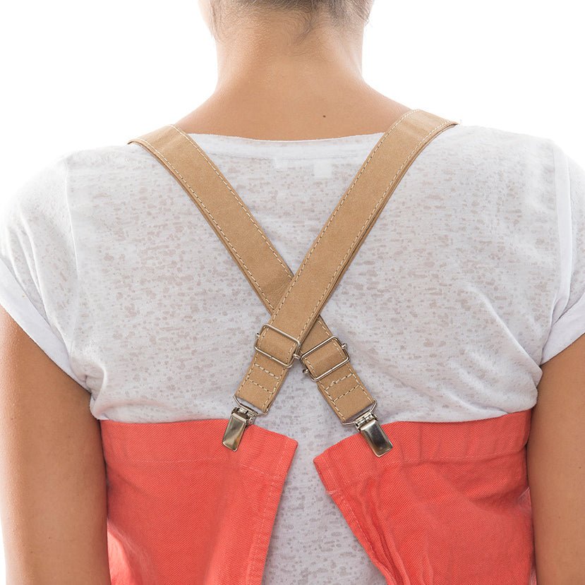 A woman is shown wearing a white t-shirt, from the back. She wears a watermelon coloured washable paper apron with crossed washable paper straps in tan. 
