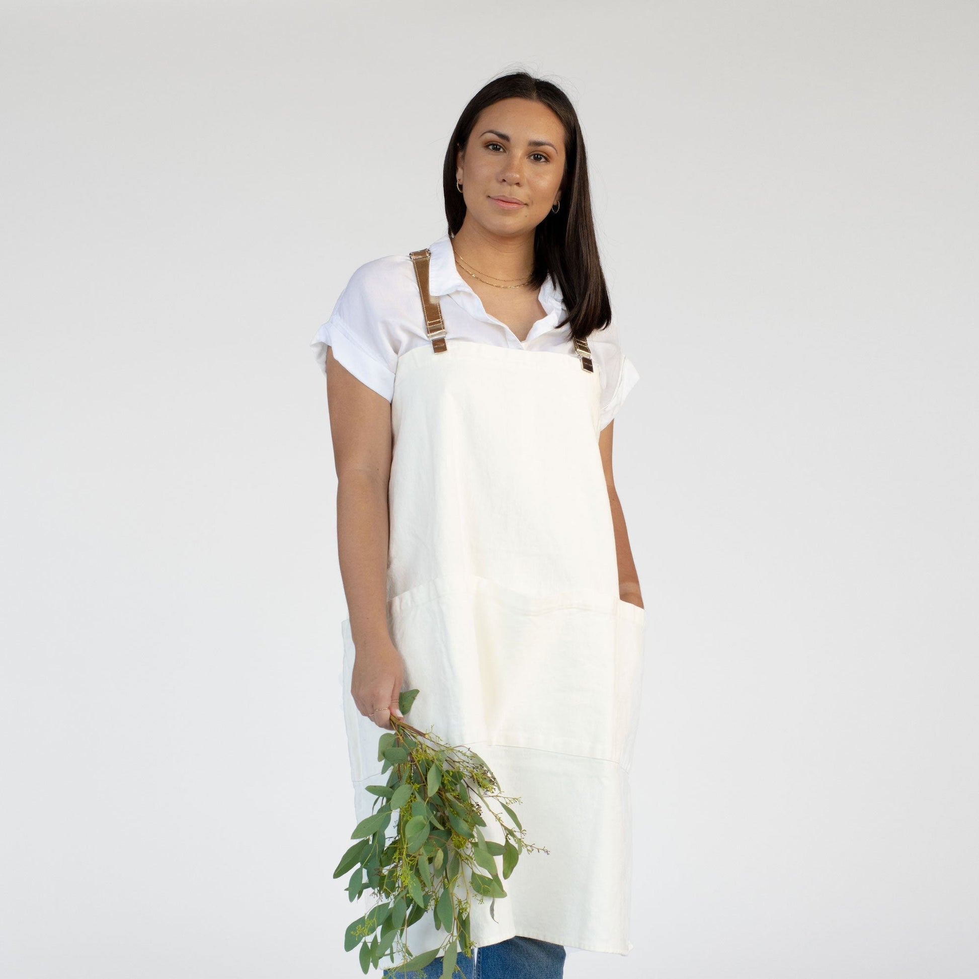 A woman is shown from the front holding a bunch of flowers. She is wearing a white shirt and a cream washable paper apron with gold metallic washable paper straps.
