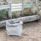 A washable paper bag is shown. The bag is rolled down at the top and features a UASHMAMA logo label on the bottom left corner. The bag pictured is the gigantic size in pale grey. The bag is shown sitting on a beach with a driftwood log behind it and a painted wooden fence.