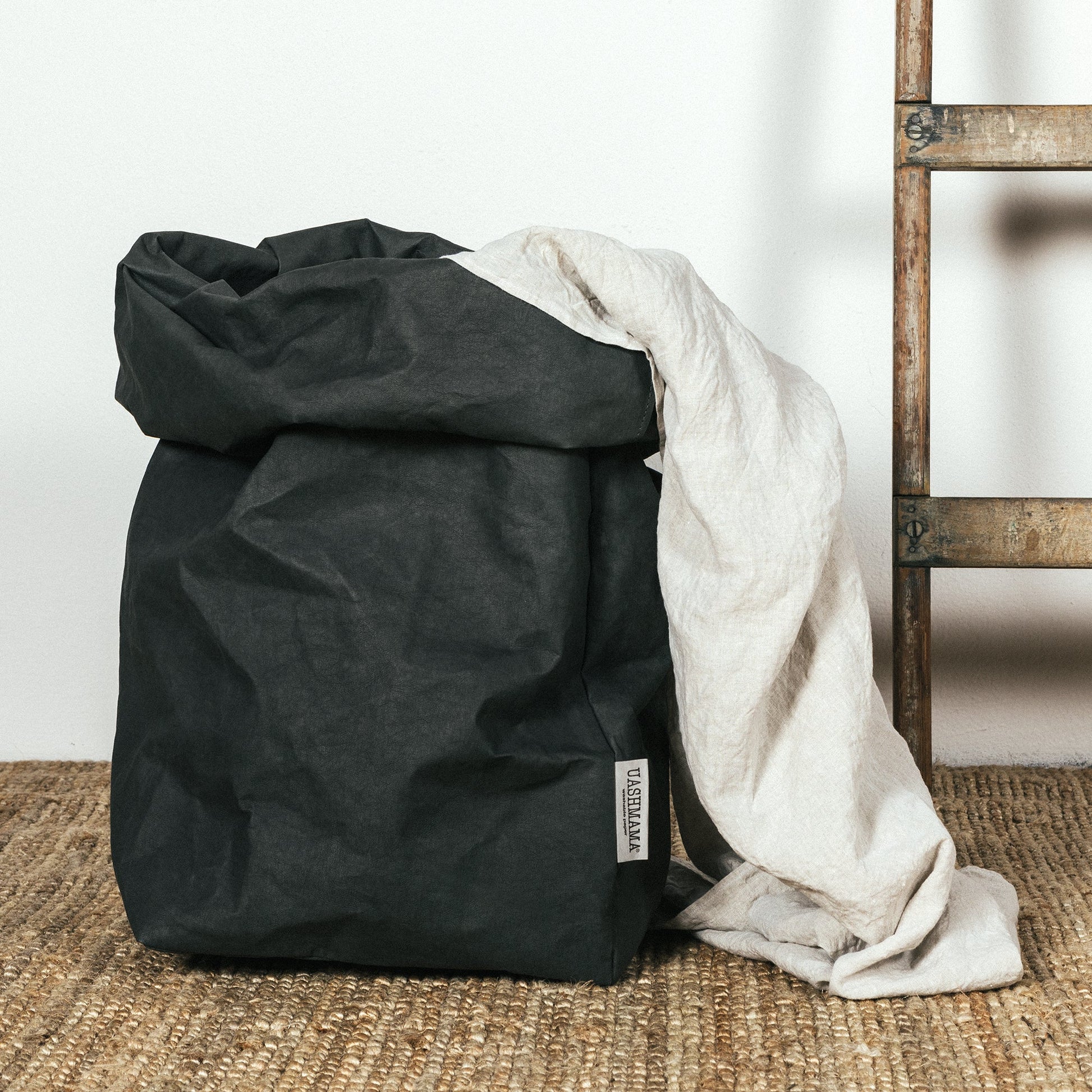 A washable paper bag is shown. The bag is rolled down at the top and features a UASHMAMA logo label on the bottom left corner. The bag pictured is the gigantic size in black. There is a blanket shown draped out of the bag, and next to the blanket and bag is a wooden ladder.