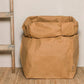 A washable paper bag is shown. The bag is rolled down at the top and features a UASHMAMA logo label on the bottom left corner. The bag pictured is the gigantic size in tan. Next to the washable paper bag is a wooden ladder.