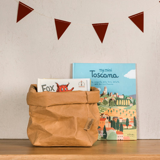 A washable paper bag is shown. The bag is rolled down at the top and features a UASHMAMA logo label on the bottom left corner. The bag pictured is the large plus size in tan. The bag is shown holding a child's book. Behind the bag, a children's book on Tuscany is leaning against the wall. Hanging on the wall is some red triangular washable paper bunting.