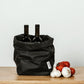 A washable paper bag is shown. The bag is rolled down at the top and features a UASHMAMA logo label on the bottom left corner. The bag pictured is the large plus size in black. Two wine bottles are shown inside the bag. To the right of the bag are three tomatoes and two bulbs of garlic.