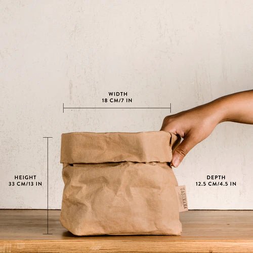A washable paper bag is shown. The bag is rolled down at the top and features a UASHMAMA logo label on the bottom left corner. The bag pictured is the medium size in tan. The dimensions are shown on the image and a hand is shown holding the side of the bag.