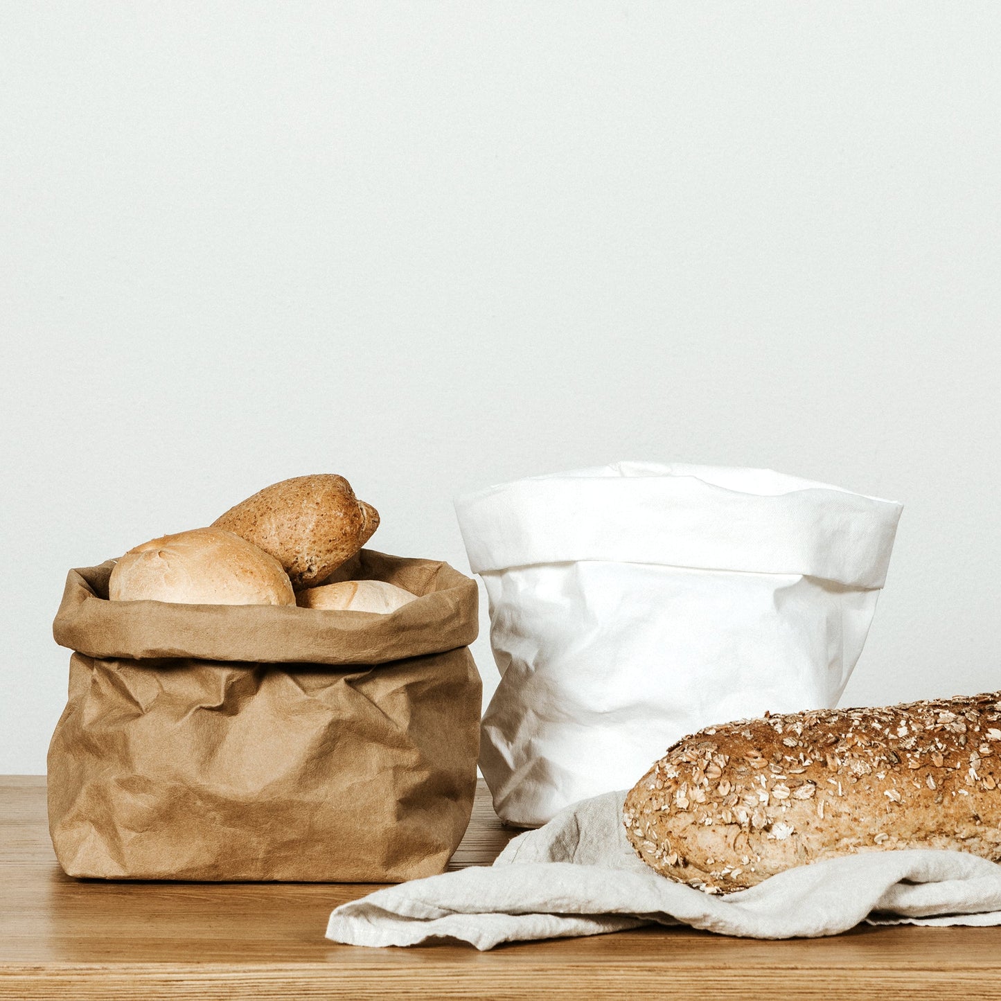 A washable paper bag is shown. The bag is rolled down at the top and features a UASHMAMA logo label on the bottom left corner. The bag pictured is the medium size in tan. The tan paper bag is filled with bread rolls. Next to it is a medium washable paper bag in white which is empty. Also shown is a loaf of seeded bread lying on a linen napkin.