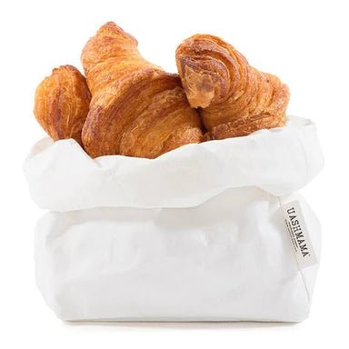A washable paper bag is shown. The bag is rolled down at the top and features a UASHMAMA logo label on the bottom left corner. The bag pictured is the medium size in white. It is shown filled with croissants.