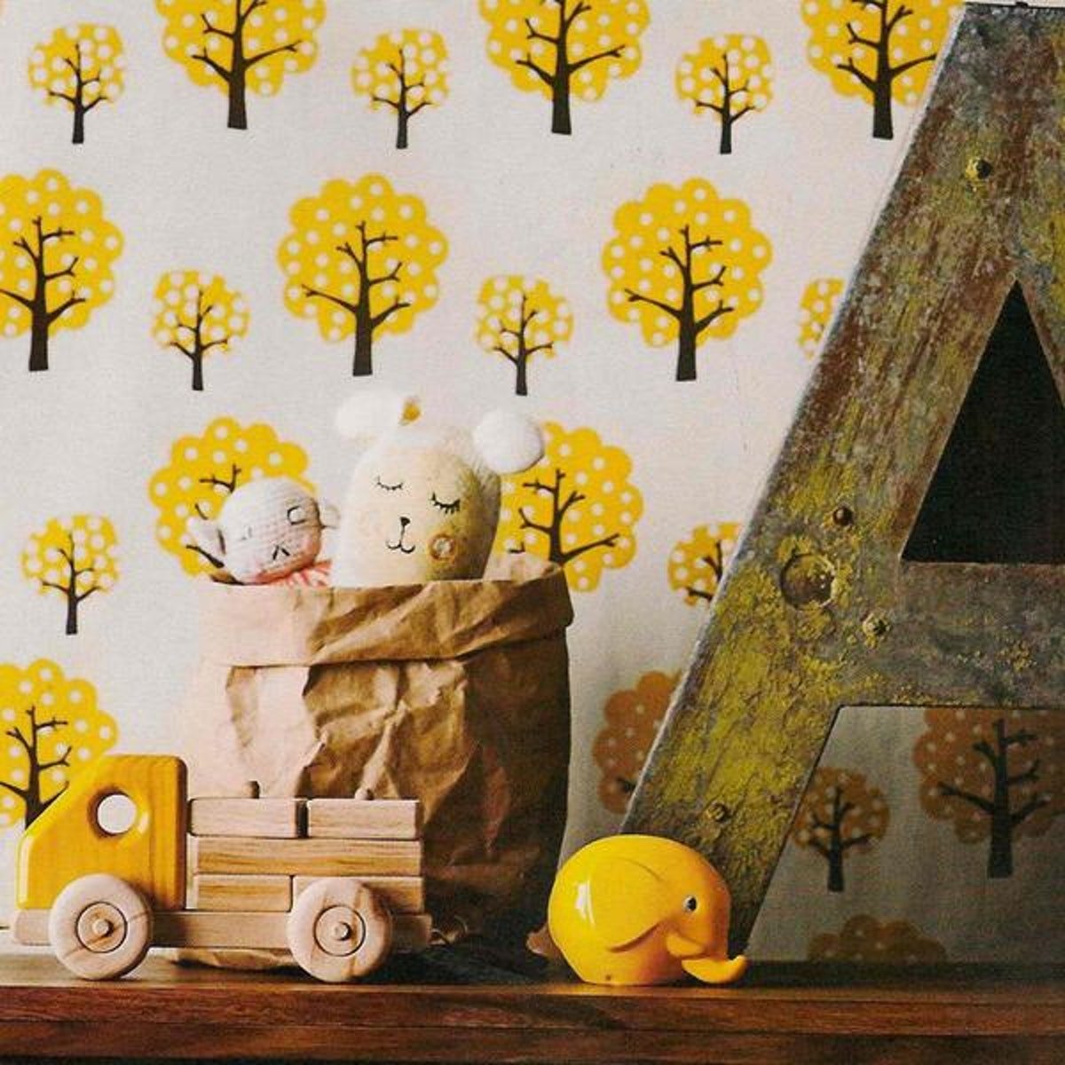A wooden shelf is shown in front of a wall papered in wallpaper with yellow trees. On the shelf is a washable paper bag in tan in the medium size, containing two soft toys. In front of the paper bag is a wooden truck toy and a small yellow elephant toy. Next to the paper bag is a large wooden A letter. 
