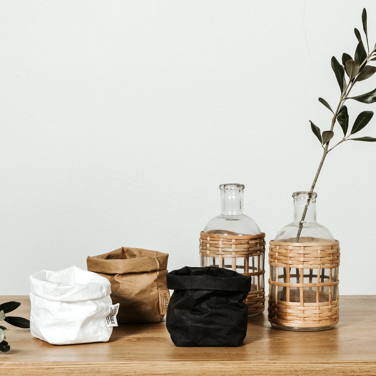 Three piccolo washable paper bags are shown. From left to right they are white, tan and black in colour and are all empty. Next to the black bag on the left are two glass vases with raffia. A tall green stem is in the vase on the right.