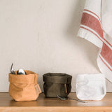 Three piccolo washable paper bags are shown. The bag on the left is tan and is filled with teaspoons. Next on the right are a dark green and a white washable paper bag which are both empty. In front of them lies a teaspoon. Draped in the background is a linen tea towel.