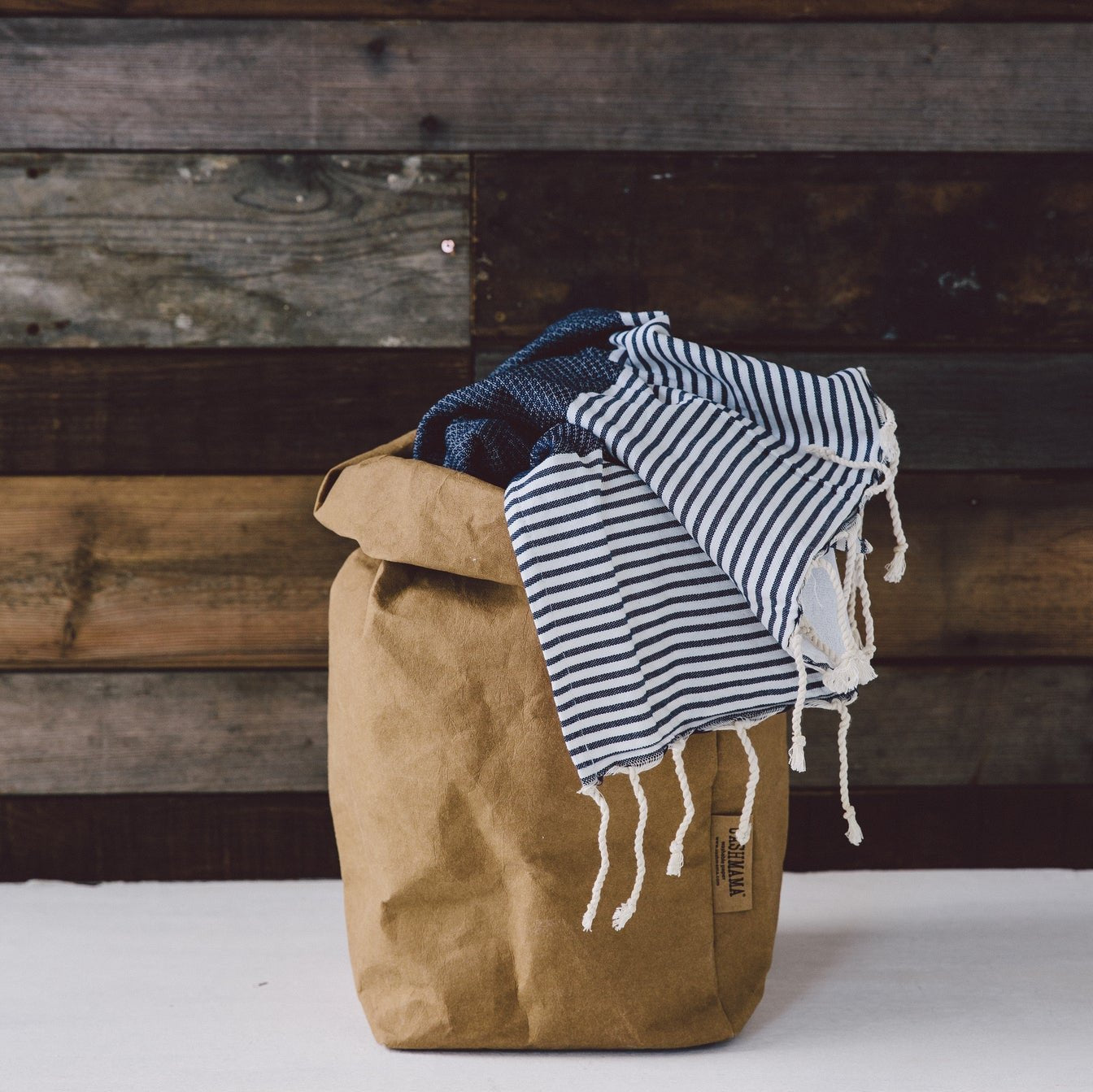 A washable paper bag is shown. The bag is rolled down at the top and features a UASHMAMA logo label on the bottom left corner. The bag pictured is the extra large size in tan. Inside the paper bag is a navy and white striped blanket.