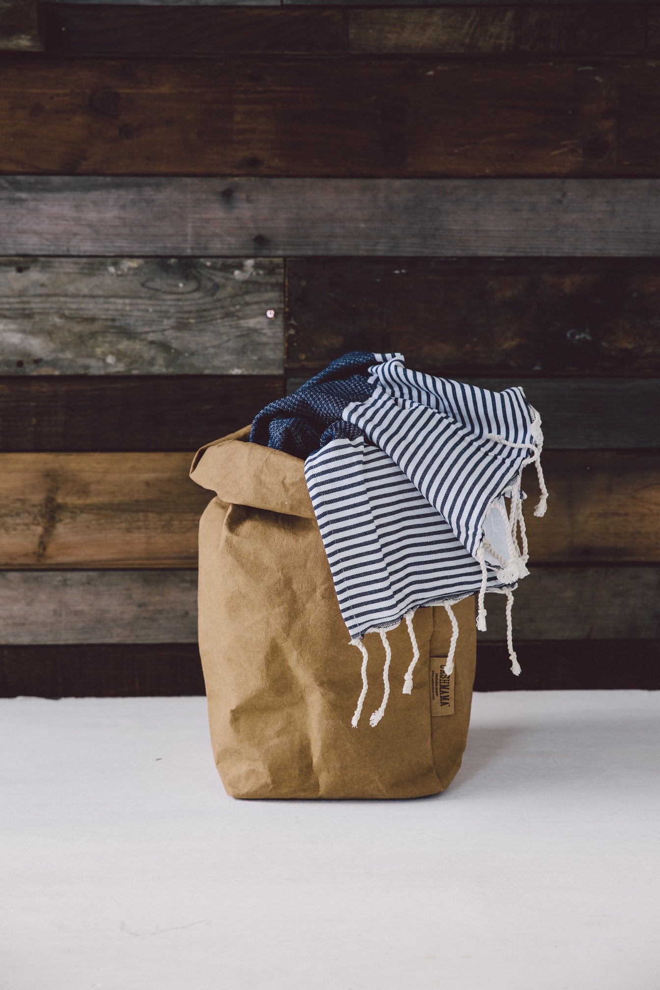 A washable paper bag is shown. The bag is rolled down at the top and features a UASHMAMA logo label on the bottom left corner. The bag pictured is the extra large size in tan. Inside the paper bag is a navy and white striped blanket.