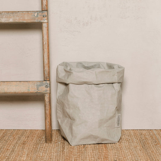 A washable paper bag is shown. The bag is rolled down at the top and features a UASHMAMA logo label on the bottom left corner. The bag pictured is the extra large size in pale grey. Next to the paper bag is a wooden ladder.