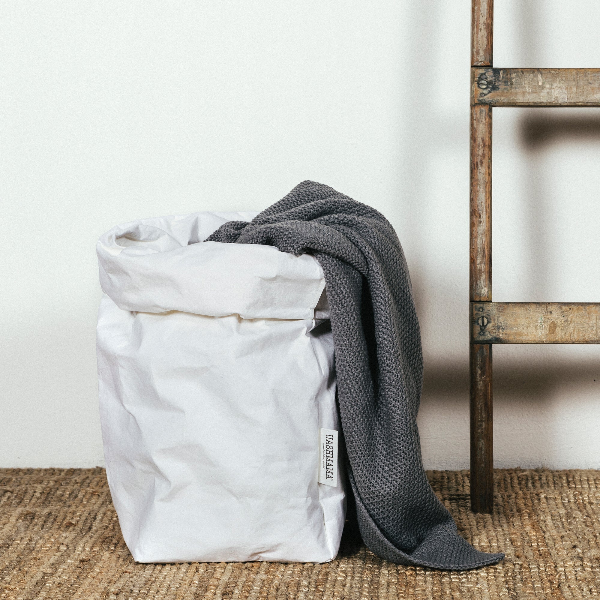 A washable paper bag is shown. The bag is rolled down at the top and features a UASHMAMA logo label on the bottom left corner. The bag pictured is the extra large size in white. The bag is shown with a grey blanket draped inside it. Next to the paper bag is a wooden ladder.