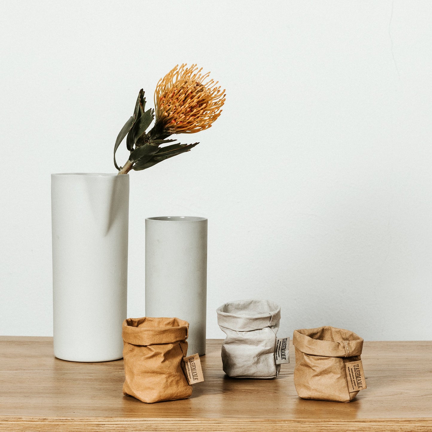 Three extra small washable paper bags are shown sitting on a wooden surface. Two bags are tan and one is pale grey. To the right of the image are two cylindrical vases in pale grey. One vase is holding a dried flower.