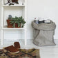 An extra extra large paper bag is shown in grey containing rolled up posters. Next to the  washable paper bag is a white shelving unit displaying plants. In front is a patterned rug and a pair of brown clogs.