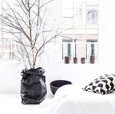 An entirely white room is shown. On the left is an extra extra large washable paper bag in metallic black containing a tree. The branches are bare. Next to the bag on the floor is a black wooden bowl. A bed with white bedding and a black and white cushion is also shown.