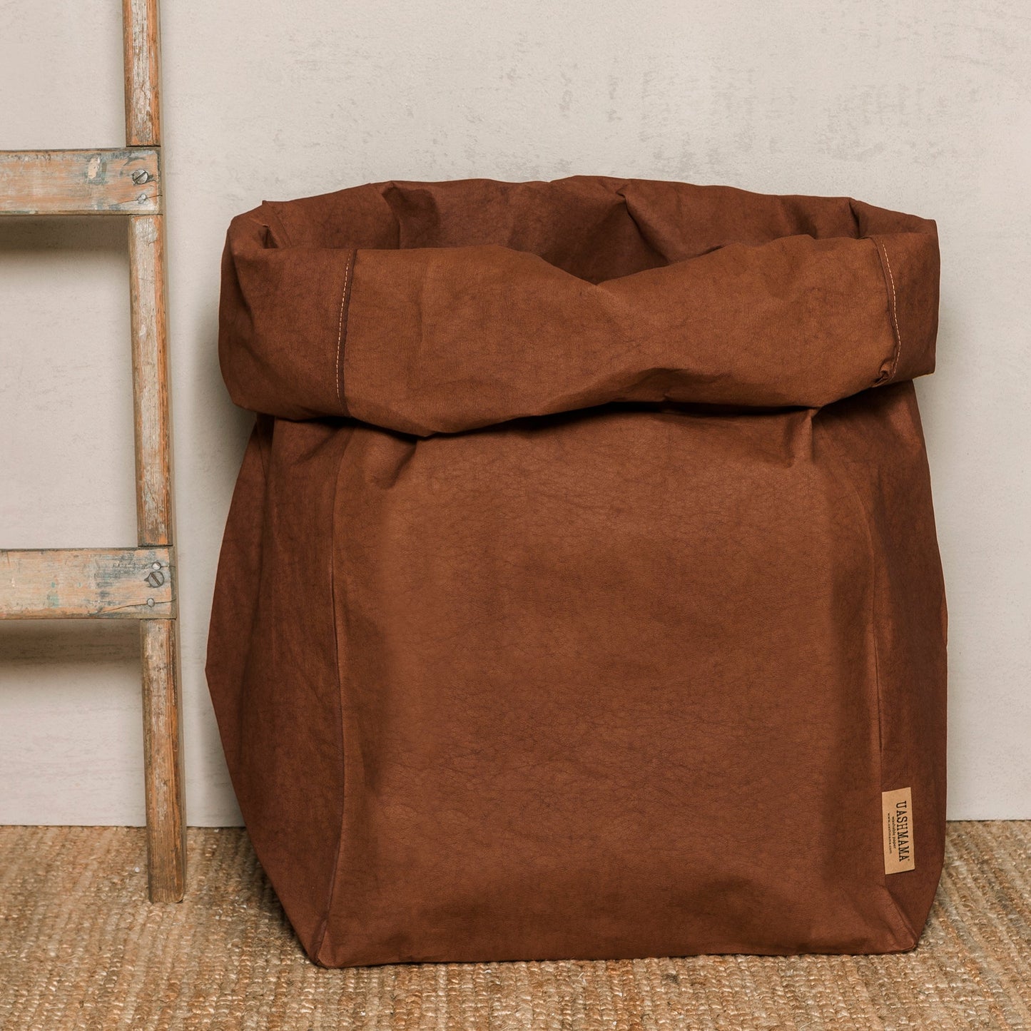 A washable paper bag is shown. The bag is rolled down at the top and features a UASHMAMA logo label on the bottom left corner. The bag pictured is the gigantic size in brown. Next to the paper bag is a wooden ladder.
