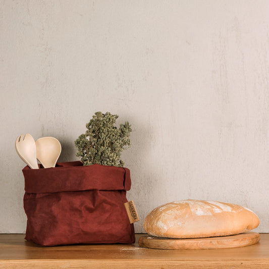 A washable paper bag is shown. The bag is rolled down at the top and features a UASHMAMA logo label on the bottom left corner. The bag pictured is the large size in a brown colour. Inside the bag are some herbs and wooden salad servers. Next to the bag isa loaf of bread on a wooden bread board.