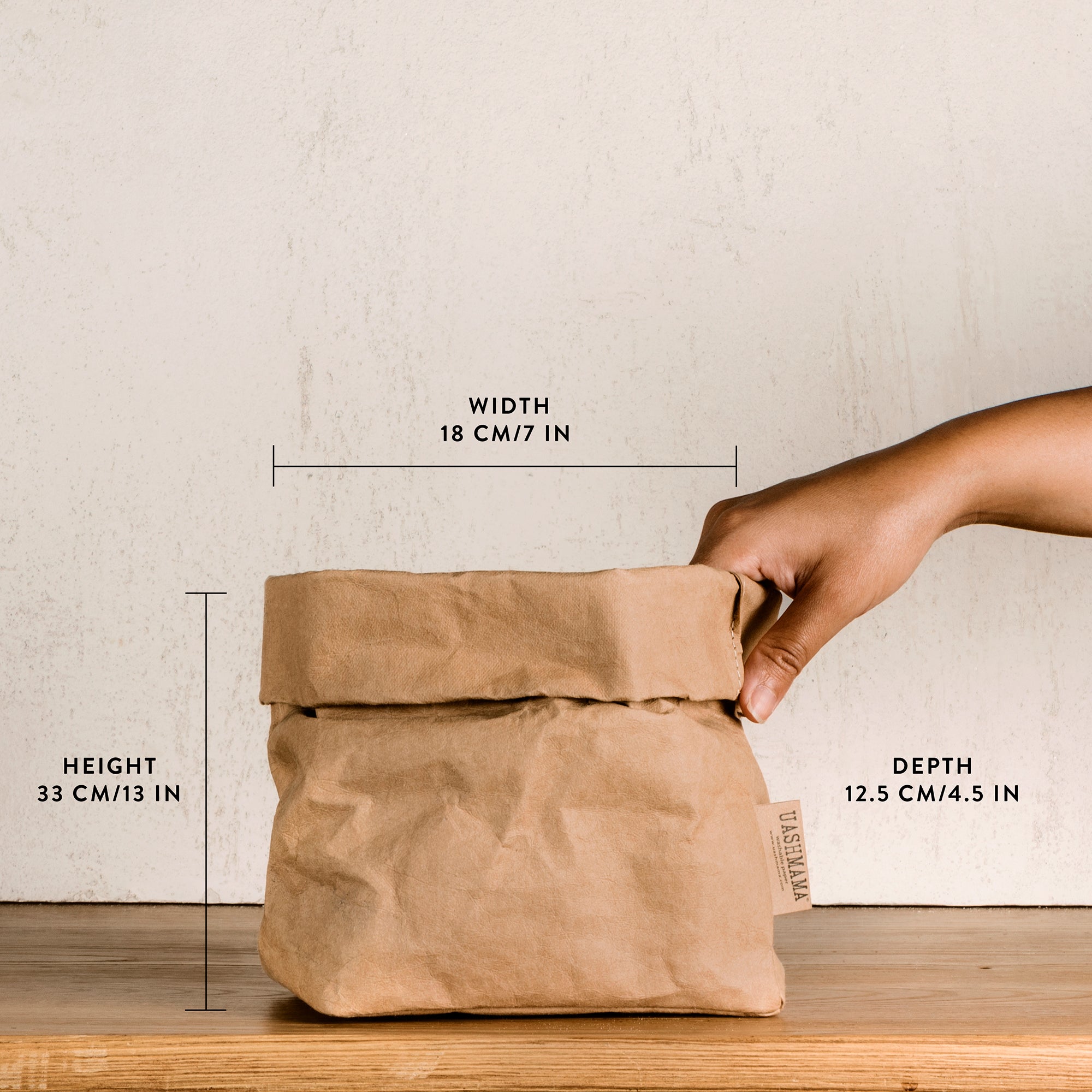 A washable paper bag is shown. The bag is rolled down at the top and features a UASHMAMA logo label on the bottom left corner. The bag pictured is the medium size in tan. The dimensions are shown in the image and a hand is shown holding the right side of the bag.