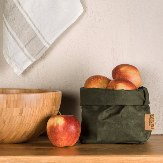 A washable paper bag is shown. The bag is rolled down at the top and features a UASHMAMA logo label on the bottom left corner. The bag pictured is the medium size in dark green. The paper bag is filled with apples. To the left of the bag is a wooden bowl and a red apple. In the background a linen tea towel is shown draped on the wall.