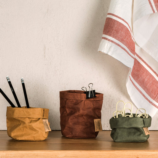 Three piccolo washable paper bags are shown. On the left is a tan washable paper bag with three pencils. In the middle is a brown washable paper bag with a binder clip clipped to the top. On the right is a dark green washable paper bag filled with large paper clips. On the wall behind a linen tea towel is draped.
