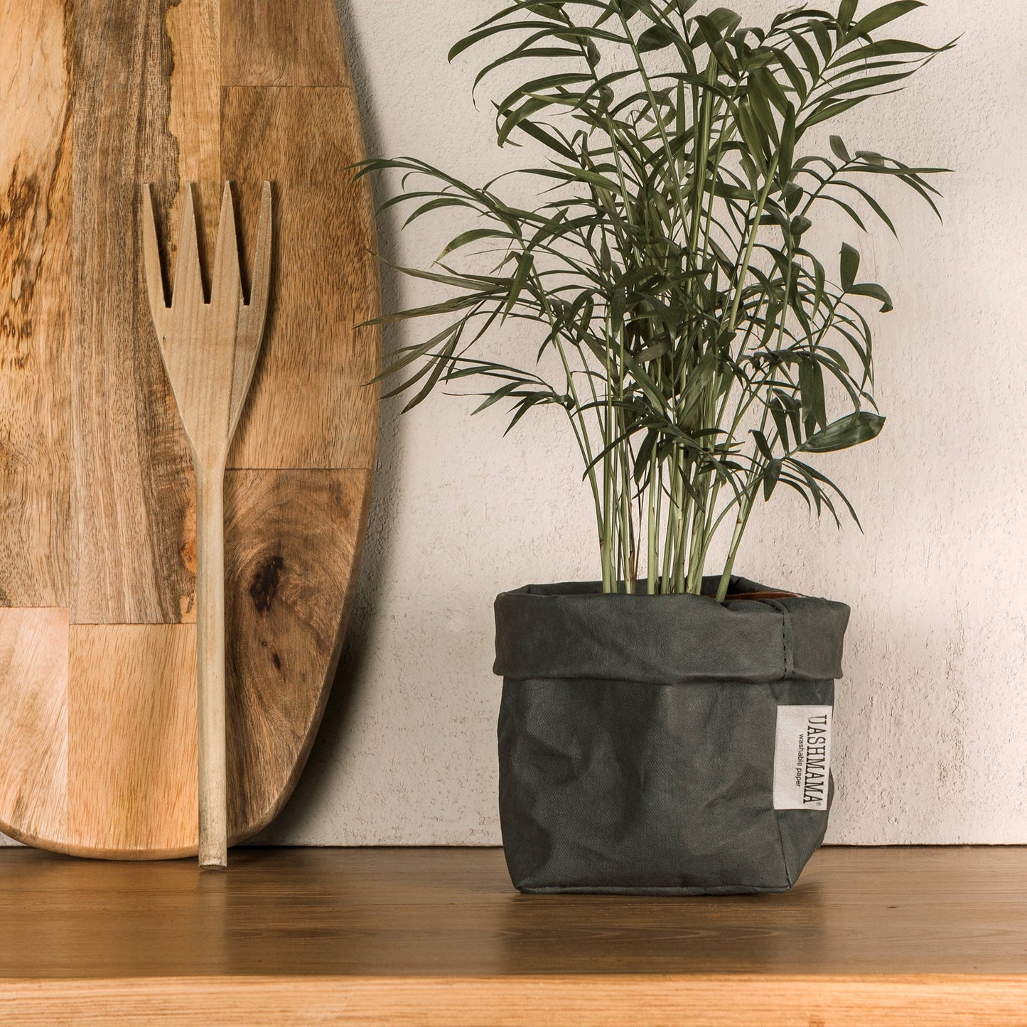 A small dark grey washable paper bag is shown containing a tall green leafy plant. On the left leaning against the wall are a wooden chopping boar and wooden fork.