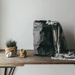 A set of three washable paper bags are displayed on a wooden table. On the left a small grey washable paper bag contains a succulent plan. To the right of that is an extra small washable paper bag containing a very small succulent plant. To the right of them both is an extra large dark grey washable paper bag. There is a blanket spilling out of the paper bag. The blanket is light grey with a dark grey pattern.