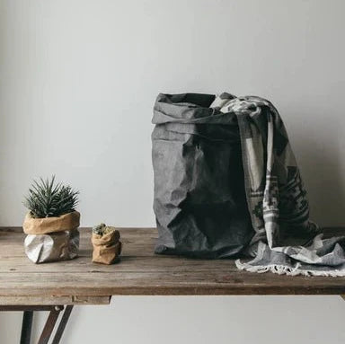 A set of three washable paper bags are displayed on a wooden table. On the left a small grey washable paper bag contains a succulent plan. To the right of that is an extra small washable paper bag containing a very small succulent plant. To the right of them both is an extra large dark grey washable paper bag. There is a blanket spilling out of the paper bag. The blanket is light grey with a dark grey pattern.