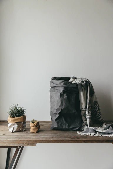 A set of three washable paper bags are displayed on a wooden table. On the left a small grey washable paper bag contains a succulent plan. To the right of that is an extra small washable paper bag containing a very small succulent plant. To the right of them both is an extra large dark grey washable paper bag. There is a blanket spilling out of the paper bag. The blanket is light grey with a dark grey pattern.