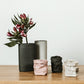 Three extra small washable paper bags are on a wooden surface. Each bag is empty. From left to right they are pale pink, black and pale cream in colour. To the left of the washable paper bags are two vases. The black vase on the left contains two stems of flowers.